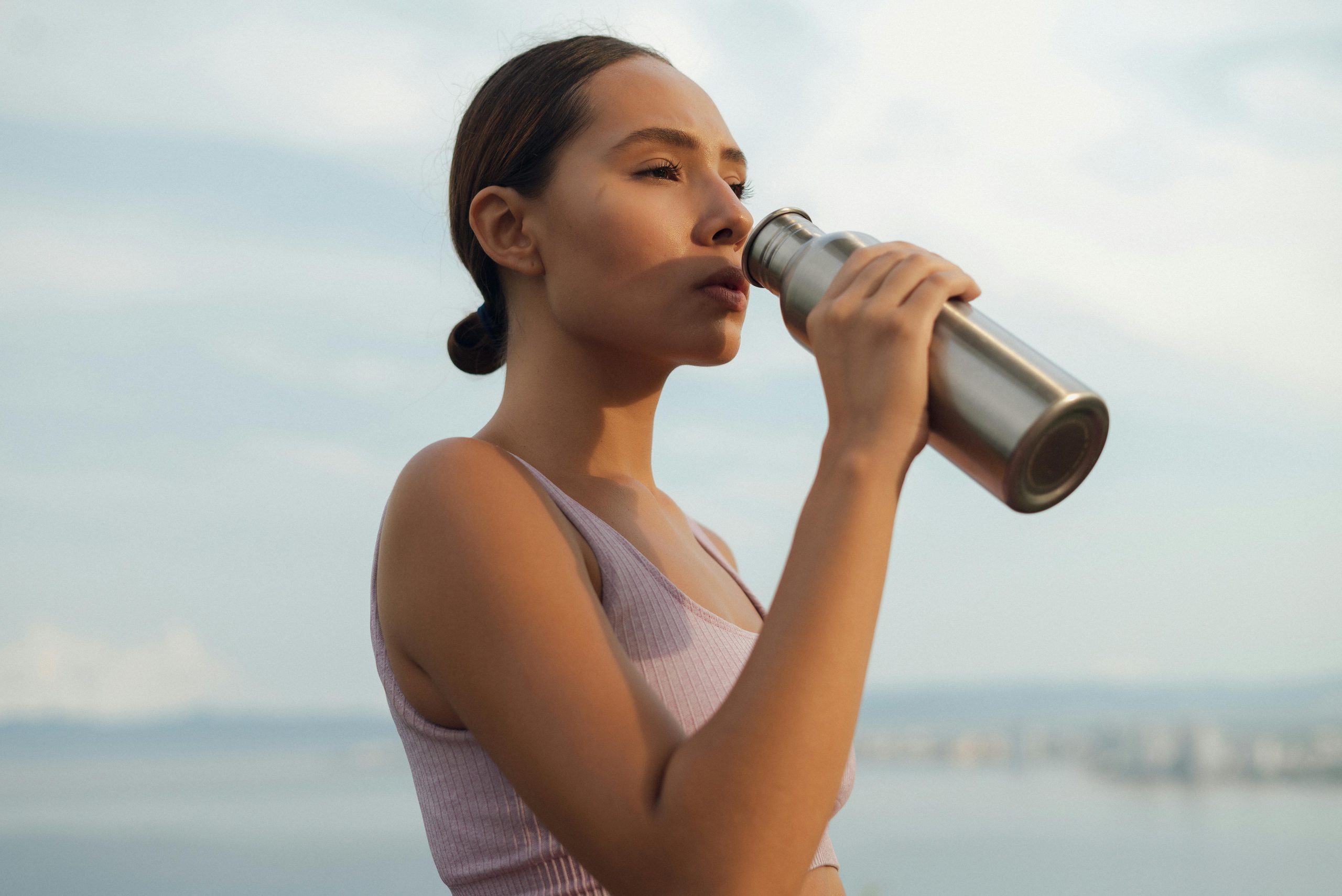 A woman drinks from a metal bottle on a sunny day by the beach, wearing a sports bra.